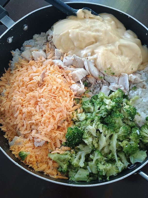 Chicken broccoli rice casserole ingredients combined in a skillet before baking. Shredded cheddar cheese chopped chicken, broccoli, rice, and creamy sauce are arranged together in the pan.