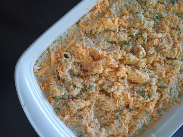 Unbaked chicken broccoli rice casserole spread in a white baking dish and topped with shredded cheddar cheese.