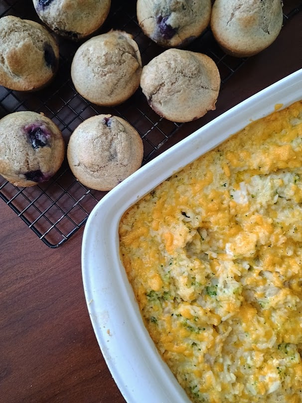 Chicken broccoli rice casserole baked with melted cheddar cheese in a white casserole dish. Fresh blueberry muffins cool on a wire rack beside it.
Image: 2