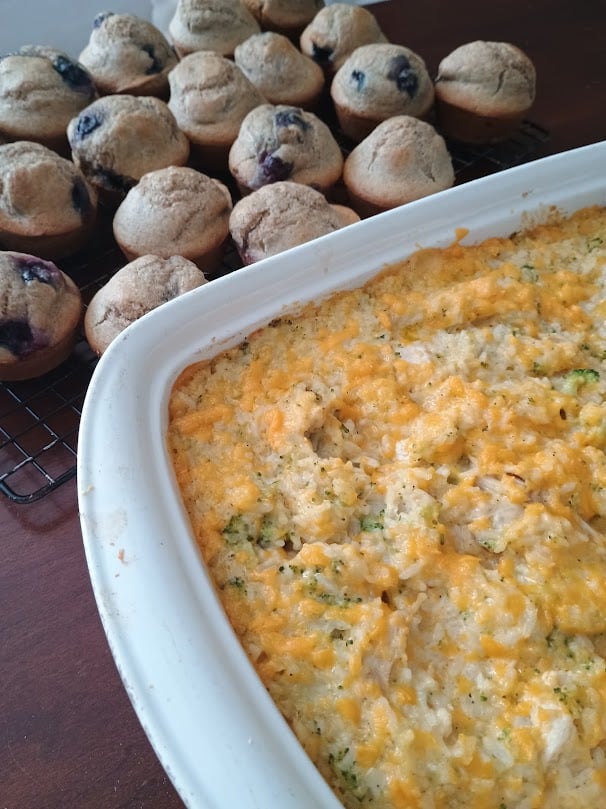 Cheesy chicken rice broccoli casserole in a white baking dish with a golden melted cheese topping. A batch of freshly baked blueberry muffins sits on a cooling rack in the background.