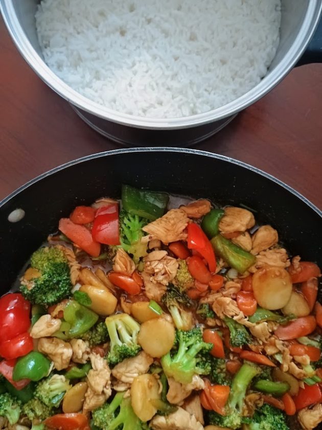 Chicken stir fry with broccoli, carrots, red and green bell peppers, chicken, and water chestnuts in a brown sauce, with a pot of cooked white rice ready for serving.