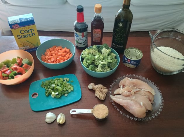 Ingredients for homemade chicken stir fry arranged on a table, including raw chicken breasts, broccoli, carrots, bell peppers, green onions, ginger, garlic, brown sugar, corn starch, soy sauce, oil, water chestnuts, and rice.