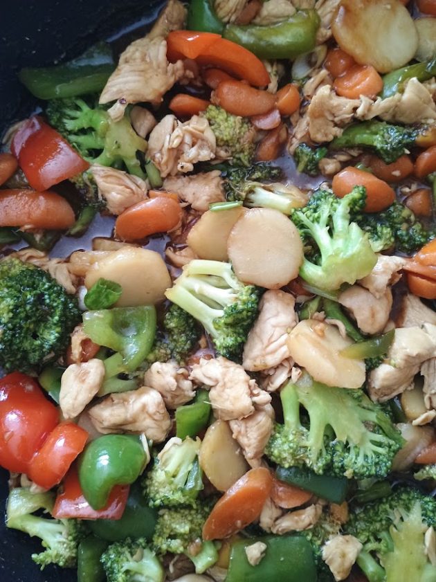 Homemade chicken stir fry in a black skillet served alongside a pot of fluffy white rice, with colorful broccoli, carrots, bell peppers, chicken, and water chestnuts.