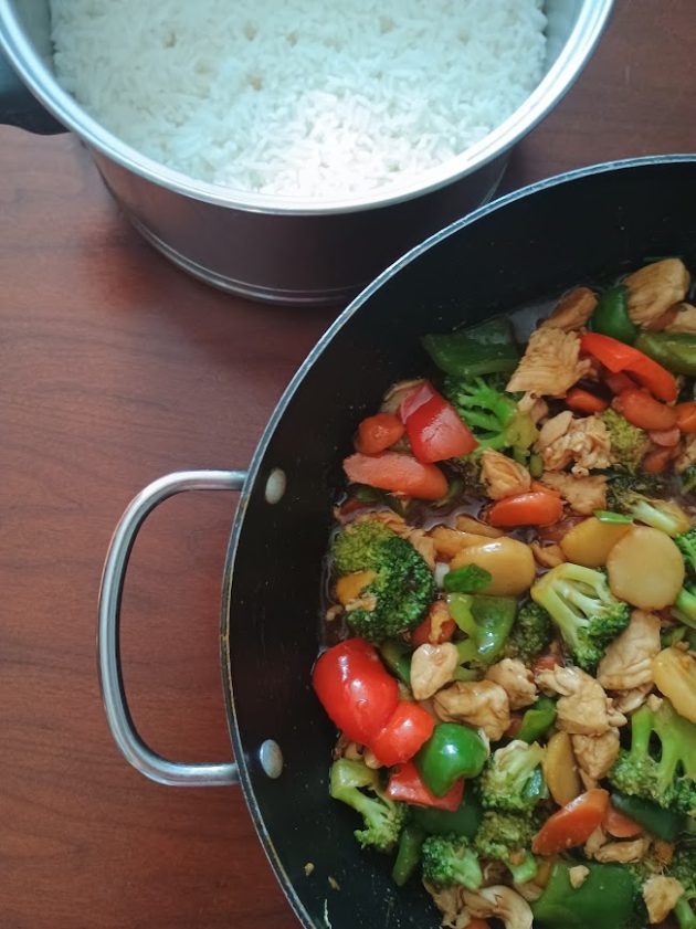 Finished chicken stir fry in a black skillet beside a pot of cooked white rice, with chicken, broccoli, carrots, red and green bell peppers, and water chestnuts coated in brown sauce.