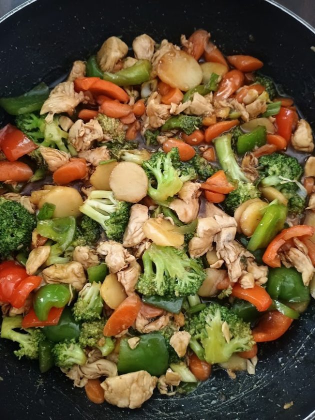 Chicken stir fry cooking in a skillet with broccoli, carrots, red and green bell peppers, water chestnuts, chicken, and green onions coated in brown sauce.