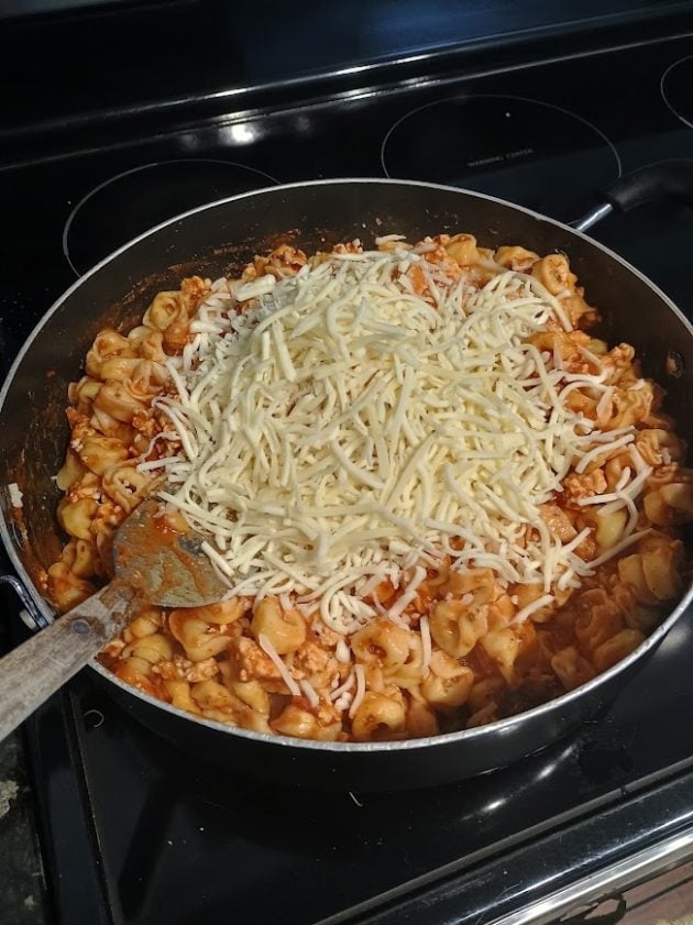 Tortellini coated in tomato meat sauce in a large skillet with a mound of shredded mozzarella cheese on top. This mixture is ready to be transferred to a dish for the baked tortellini.