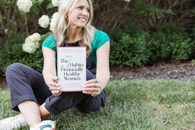 Crystal Paine sitting on the grass outdoors holding the book The 7 Habits of Financially Healthy Women.