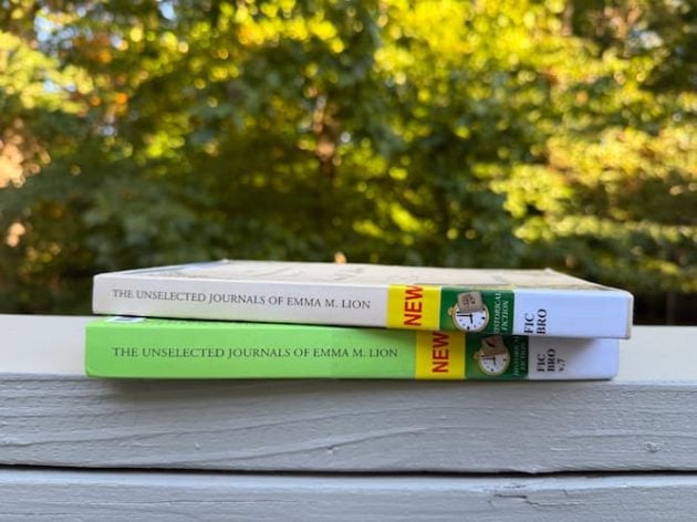 Two library copies of The Unselected Journals of Emma M. Lion stacked horizontally on a white wooden railing outdoors.