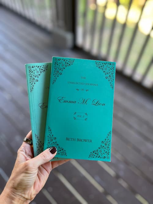 A hand holds two turquoise volumes of The Unselected Journals of Emma M. Lion by Beth Brower outdoors.