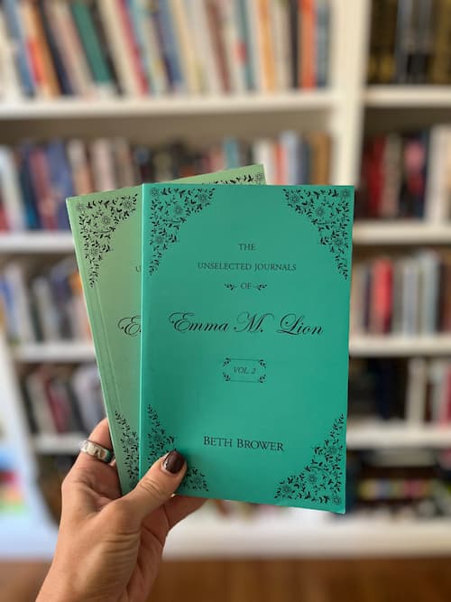 A hand holds two turquoise volumes of The Unselected Journals of Emma M. Lion by Beth Brower.